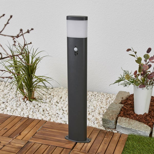 Anthrazitfarbene Außenstandleuchte mit Bewegungsmelder auf einer Holzterrasse neben weißem Zierkies und Pflanzen.