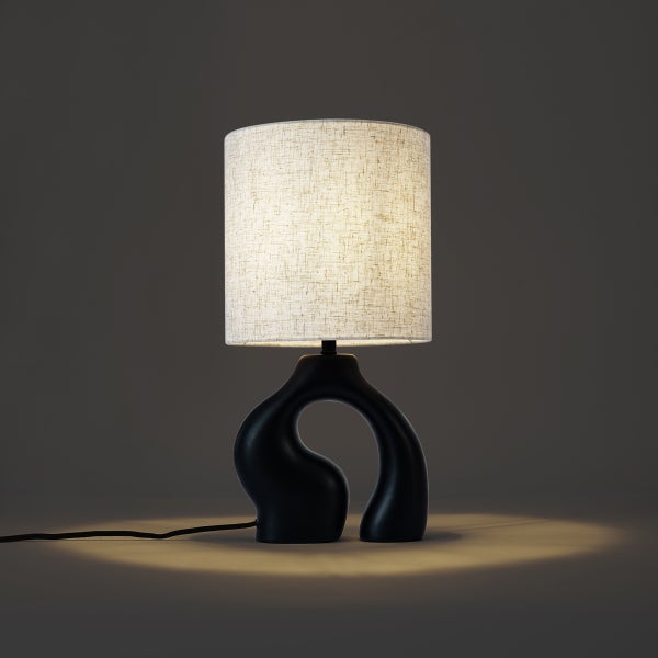 Tischleuchte mit organisch geformtem schwarzem Fuß und zylindrischem Textilschirm in Leinenoptik.