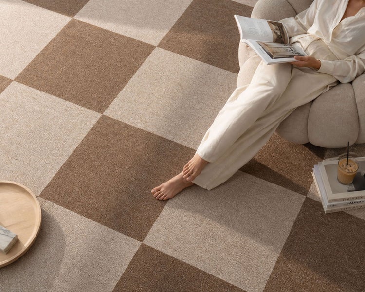 Teppichfliesen mit großem Karomuster in Braun und Beige, verlegt in einem hellen Wohnzimmer mit Sessel und einer Person, die ein Buch liest.