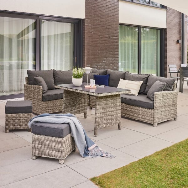Lounge-Garnitur aus Rattan-Geflecht mit Ecksofa, Esstisch und zwei Hockern auf einer Terrasse vor einer modernen Hausfassade.
