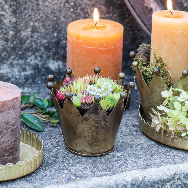 Dekorativer Metalltopf in Kronenform bepflanzt mit Hauswurz neben brennenden Stumpenkerzen auf Stein.