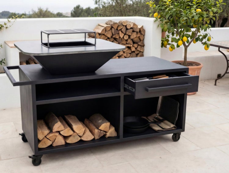 Mobiler Grillwagen aus schwarzem Metall mit integrierter Feuerschale, Grillrost, Holzlager und Schublade auf einer hellen Terrasse.