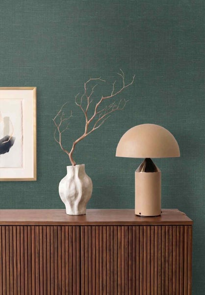 Dunkelgrüne Tapete mit Leinenstruktur hinter einem Sideboard aus Holz mit Tischleuchte und Vase.