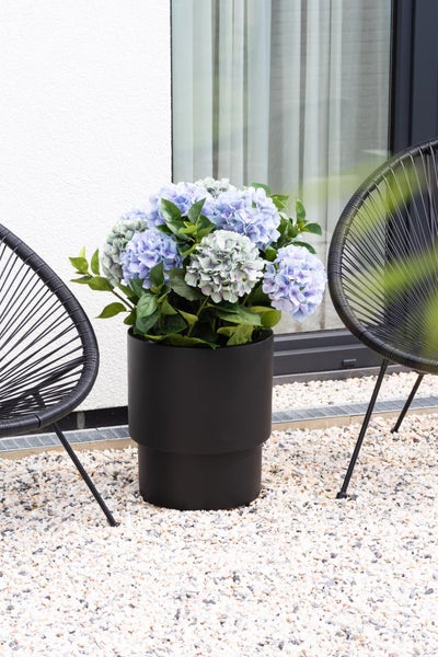 Schwarzer Pflanzkübel mit blauen Hortensien auf einer Kiesterrasse zwischen zwei schwarzen Gartenstühlen.