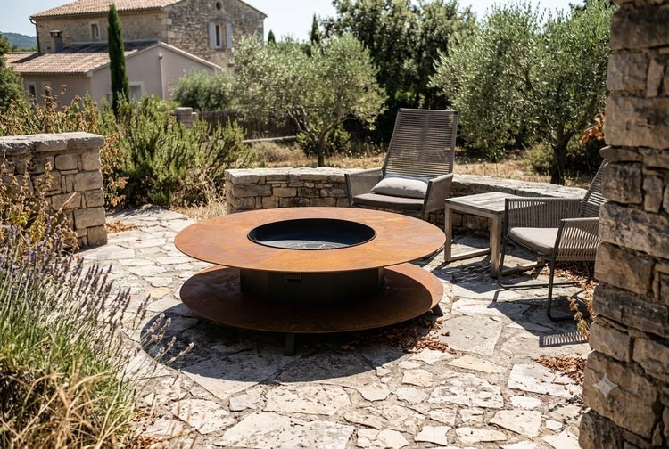 Runde Feuerstelle aus Metall in Rostoptik auf einer Steinterrasse im Garten mit zwei modernen Lounge Sesseln und einem Beistelltisch.