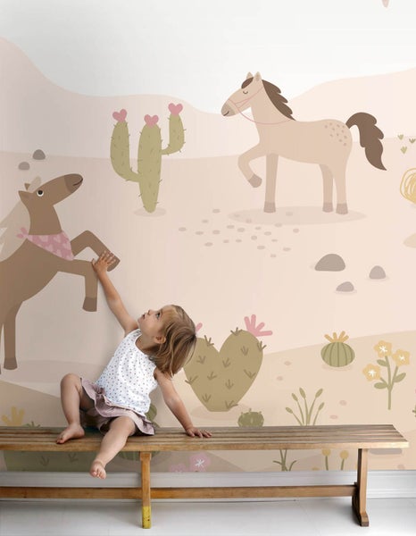 Kind auf Holzbank vor einer Wand mit Kindertapete mit Pferden, herzförmigen Kakteen und Wüstenlandschaft.