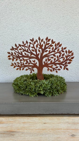 Baumfigur aus Metall mit Edelrost Patina in einem Kranz aus künstlichen grünen Blättern als Gartendekoration.