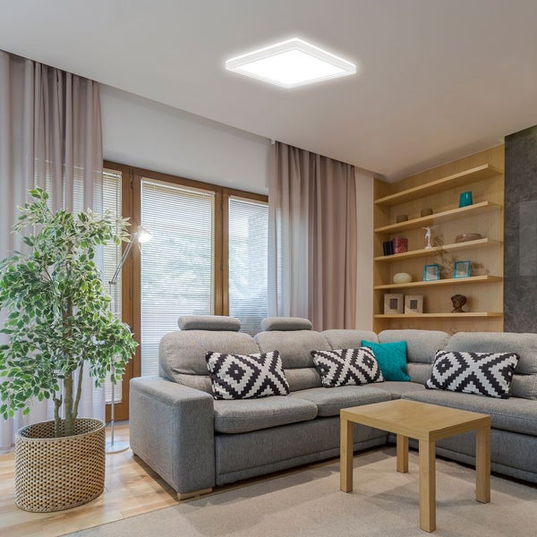 Flache, quadratische LED-Deckenleuchte in einem hellen, modernen Wohnzimmer mit Ecksofa, Holztisch und Bücherregal.