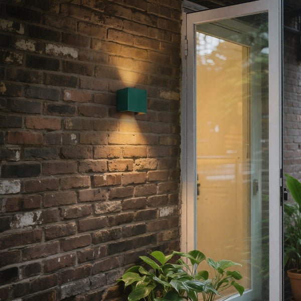 Grüne quadratische Außenwandleuchte an einer Ziegelwand neben einer Glastür, Lichtaustritt nach oben und unten.