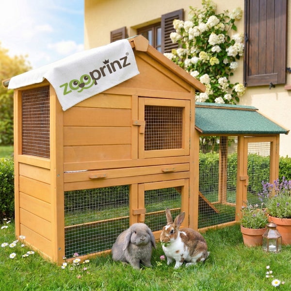 Zweistöckiger Kaninchenstall aus Holz mit integriertem Freilaufgehege und Bitumendach auf einer Rasenfläche mit zwei Kaninchen, Zooprinz Logo.