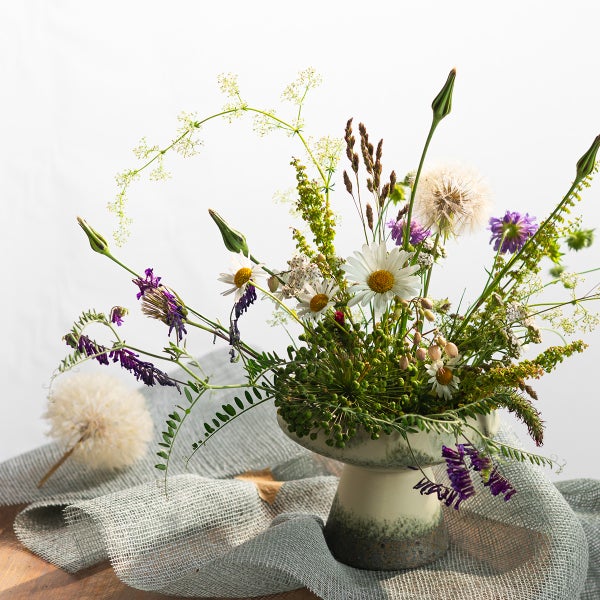 Ein Blumenstrauß aus Wiesenblumen in einer bauchigen Steingutvase auf einem strukturierten Stofftuch drapiert.