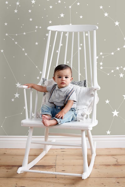 Ein Kind sitzt in einem weißen Schaukelstuhl vor einer Wandtapete mit Sternenmuster.