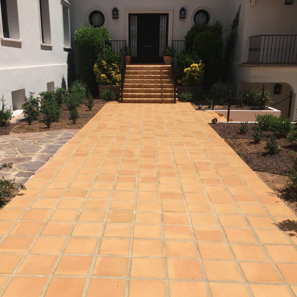 Ein gepflasterter Weg aus quadratischen Bodenfliesen führt zu einer Eingangstreppe vor einem Haus mit mediterranem Vorgarten.