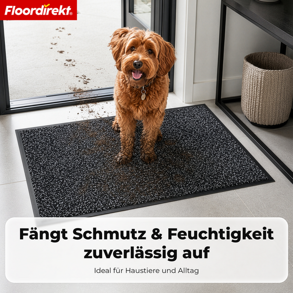 Ein mittelgroßer Hund mit schmutzigen Pfoten steht auf einer dunkelgrauen, gesprenkelten Schmutzfangmatte vor einer Glastür im Innenbereich.