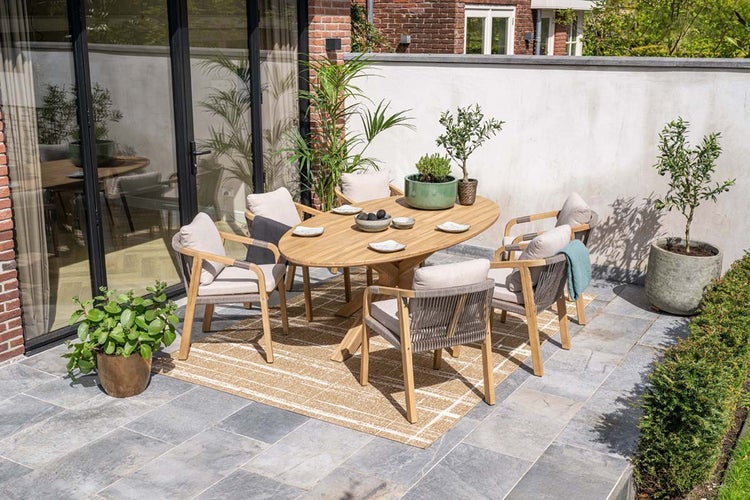 Gartenmöbel-Set mit ovalem Holztisch und sechs Holzstühlen mit Kissen auf einem gemusterten Teppich auf einer hellen, organisierten Terrasse.