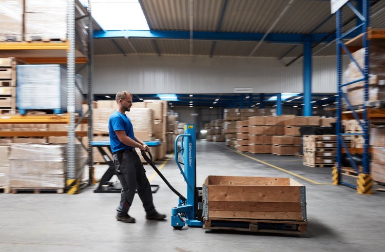 Mitarbeiter bewegt einen manuellen Hochhubwagen mit Holzpalette und Holzrahmen in einem organisierten Lager mit hohen Regalen.