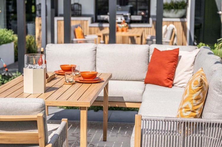 Garten-Lounge-Set aus Holz mit Ecksofa, hellen Polstern, Esstisch mit Gedeck und Tischfeuer auf einer Terrasse.