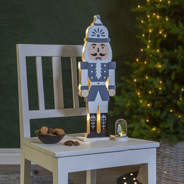 Dekorativer Nussknacker aus Holz mit LED-Beleuchtung auf einem Stuhl neben einer Schale mit Nüssen und einer Schneekugel vor einem Weihnachtsbaum