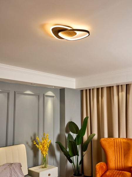 Moderne LED-Deckenleuchte mit zwei ineinandergreifenden Ringen in Holzoptik in einem Wohnraum mit Sessel und Zimmerpflanze.