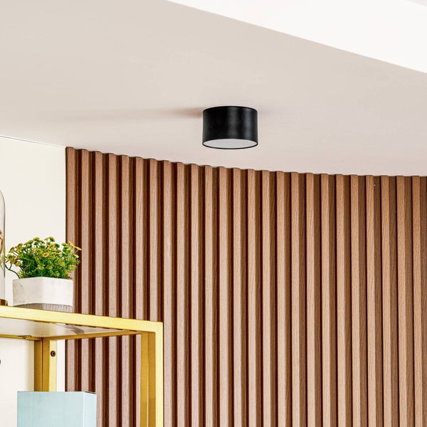 Moderne runde Deckenleuchte in Schwarz montiert an einer weißen Decke vor einer Wand mit vertikalen Holzlamellen.