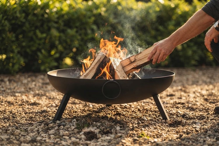 Eine runde Feuerschale aus Metall mit brennendem Holzfeuer steht auf einem Kiesboden, während eine Hand ein Holzscheit nachlegt.