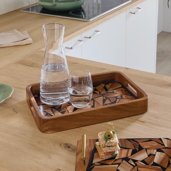 Holztablett mit geometrischem Mosaikmuster auf einer Küchenarbeitsplatte mit einer Glaskaraffe und einem Wasserglas.