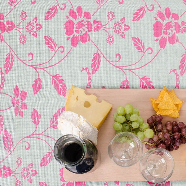 Wachstuch mit pinkem Blumenmuster, dekoriert mit einer Holzplatte mit Käse, Weintrauben und Weingläsern.
