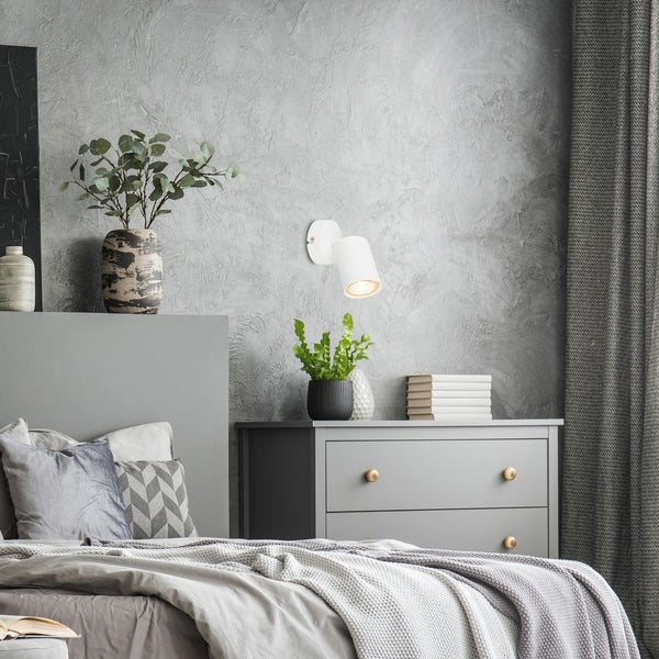 Weiße Wandleuchte über grauer Kommode in modernem, hellem Schlafzimmer mit strukturiertem Putz, Bett und Pflanzen.