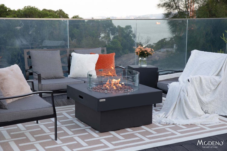 Moderne Terrasse mit quadratischer schwarzer Feuerstelle inklusive Glas-Windschutz, grauen Gartenmöbeln und einem gemusterten Außenteppich.