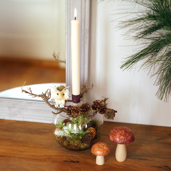 Dekorative Szene mit Kerze, Glasvase gefüllt mit Moos und Tannenzapfen, sowie zwei Pilzfiguren auf einem Holztisch.