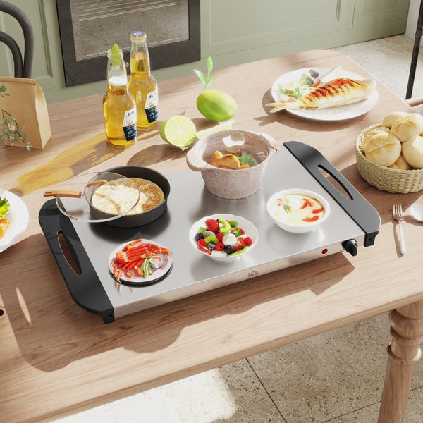 Elektrische Wärmeplatte aus Edelstahl auf einem Holztisch, bestückt mit verschiedenen Speisen in Pfannen, Töpfen und Schüsseln zum Warmhalten.