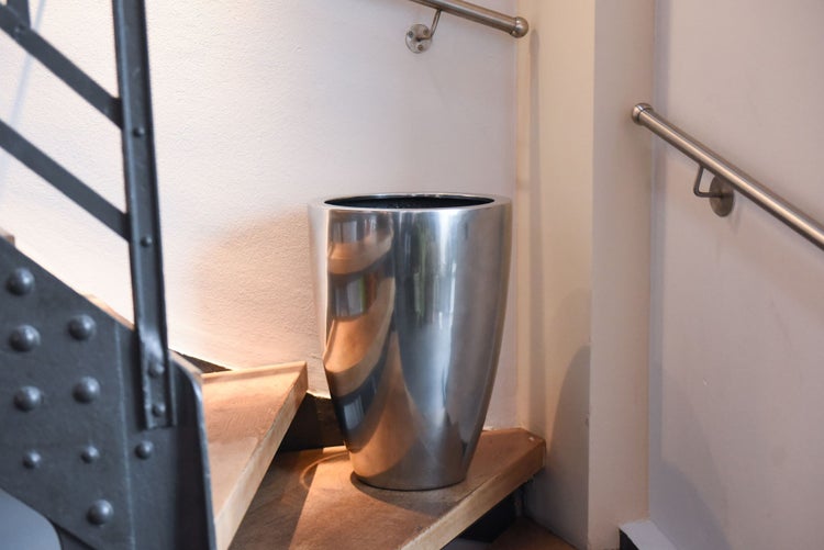 Konischer Metall-Pflanzkübel aus poliertem Edelstahl auf einem Treppenabsatz aus Holz in einem hellen Innenraum.