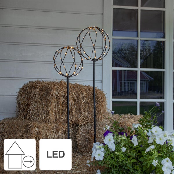 Zwei kugelförmige LED-Gartenleuchten mit Erdspieß neben einem Strohballen und Blumen
