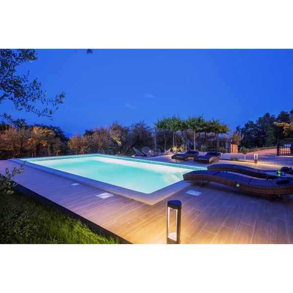 Moderner beleuchteter Außenpool bei Nacht mit Terrasse in Holzoptik, Geflecht-Sonnenliegen und dekorativer Gartenbeleuchtung.