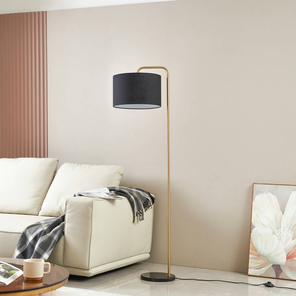 Moderne Stehleuchte mit goldfarbenem Metallgestell und schwarzem Textilschirm in einem hellen Wohnzimmer neben einem Sofa.