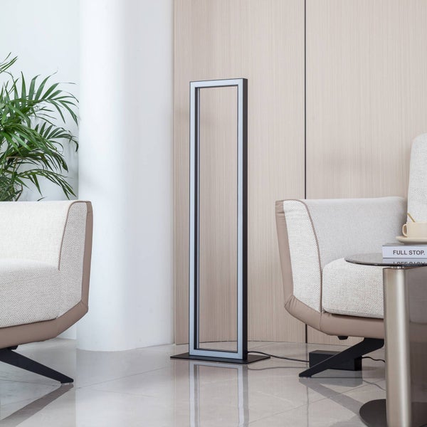 Moderne rechteckige LED-Stehleuchte aus schwarzem Metall in einem hellen Wohnzimmer zwischen zwei Sesseln vor einer hellen Holzwand.