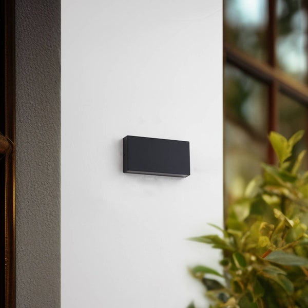 Moderne schwarze rechteckige Außenwandleuchte an einer weißen Wand neben einem Fenster und grünen Pflanzen.