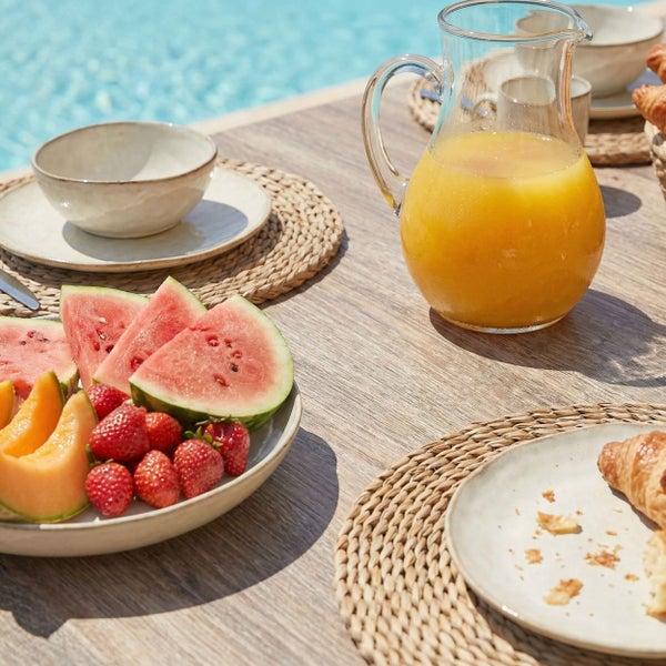 Gedeckter Holztisch am Pool mit Saftkaraffe, Obstteller mit Wassermelone und Erdbeeren sowie Keramikgeschirr auf geflochtenen Tischsets.