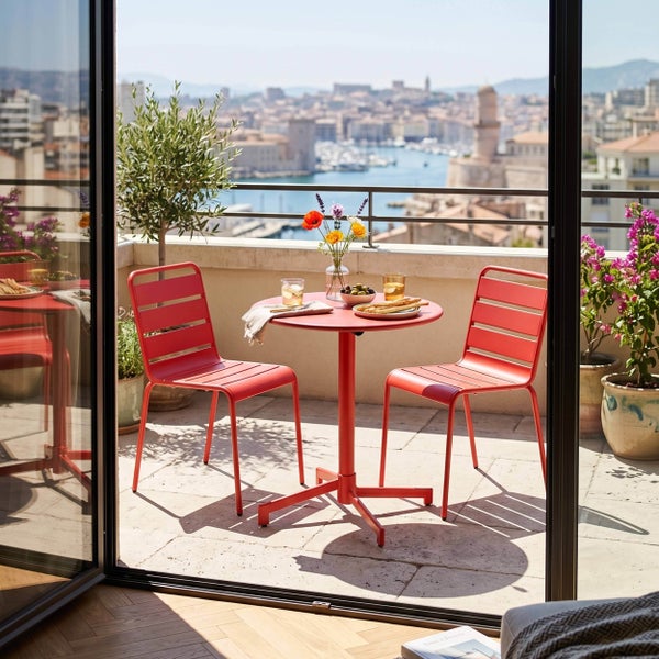 Moderner runder Balkontisch und zwei rote Metallstühle auf einem hellen Balkon mit Aussicht auf eine Stadt und einen Hafen.