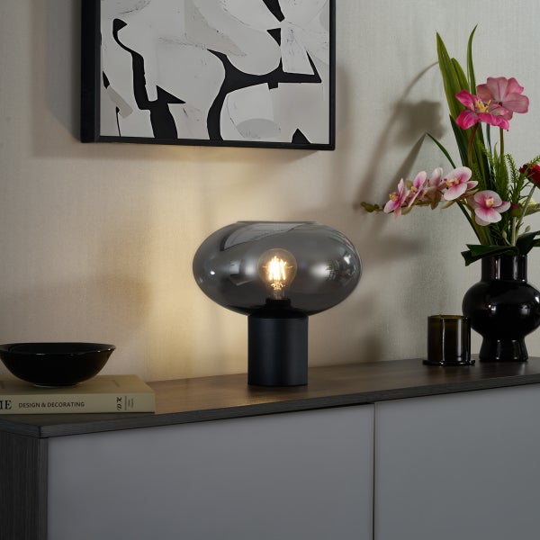 Moderne Tischleuchte mit Rauchglasschirm und schwarzem Standfuß auf einer Kommode neben einer Vase mit Orchideen und einem abstrakten Bild.