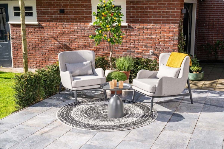 Moderne Terrasse mit zwei grauen Garten-Sesseln, rundem Beistelltisch, gemustertem Outdoor-Teppich und Zitronenbaum vor einer Backsteinwand.