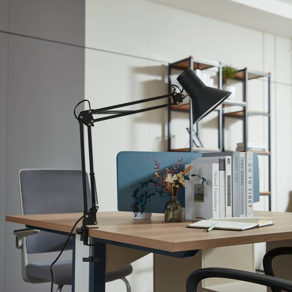 Schwarze Schreibtischleuchte mit Klemmfuß und verstellbarem Gelenkarm auf einem hölzernen Schreibtisch in einem hellen Büro.
