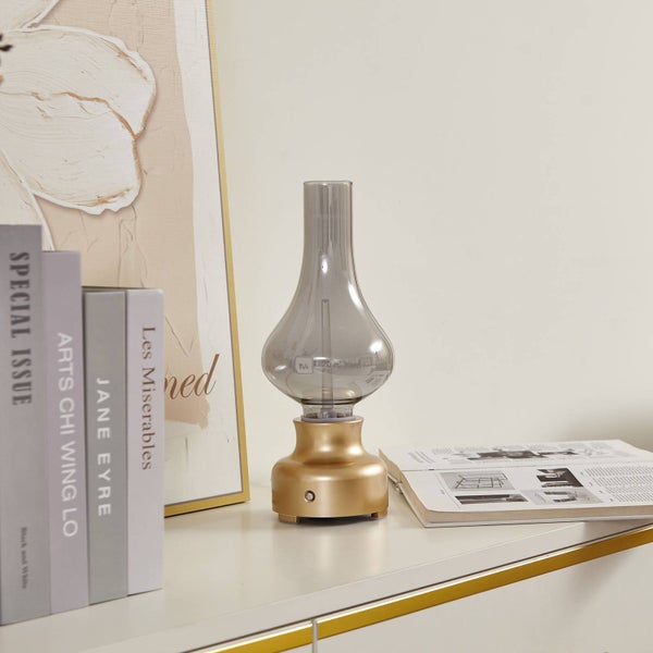 Tischleuchte im Design einer Petroleumlampe mit goldenem Fuß und rauchgrauem Glaskolben auf einem Sideboard neben Büchern.