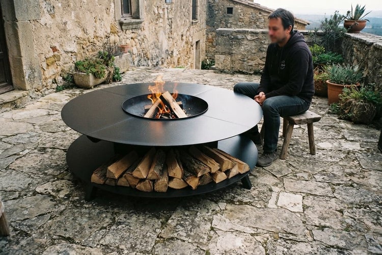 Runde Feuerstelle aus Metall mit integrierter Grillplatte und unterem Holzfach auf einer Terrasse vor einem alten Natursteinhaus