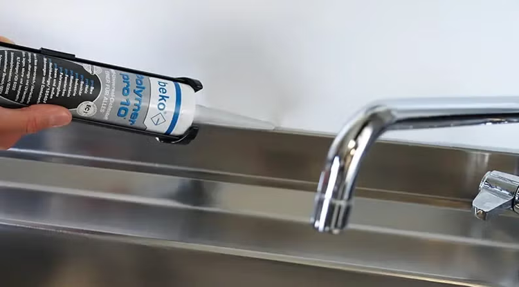 Eine Hand trägt Beko Polymer Pro 10 Dichtstoff aus einer Kartusche auf eine Spüle auf, um die Fuge zwischen Spüle und Wand abzudichten.