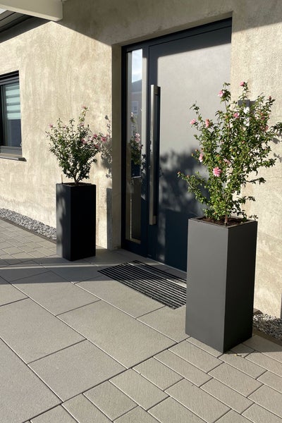 Zwei hohe quadratische Pflanzgefäße in Anthrazit mit blühenden Pflanzen vor einem modernen Hauseingang auf grauen Terrassenplatten.