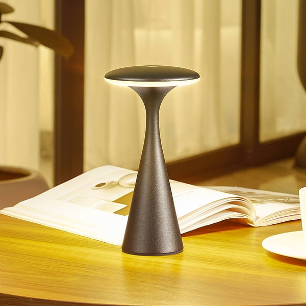 Moderne LED-Tischleuchte mit schmalem Standfuß und rundem Schirm, warmes Licht, auf einem Holztisch neben einem Buch platziert.