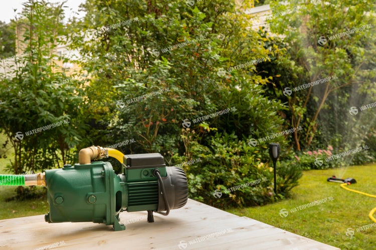 Gartenpumpe auf einer Holzoberfläche in einem Garten mit einem in Betrieb befindlichen Rasensprenger im Hintergrund.