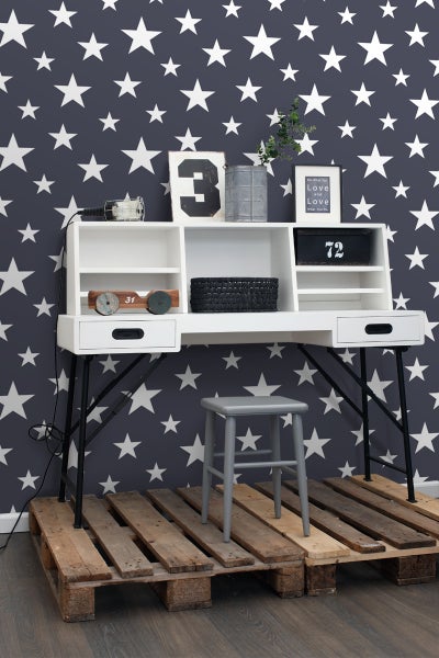 Weißer Schreibtisch mit schwarzen Beinen und grauem Hocker auf einer Holzpalette vor einer Tapete mit Sternenmuster.
