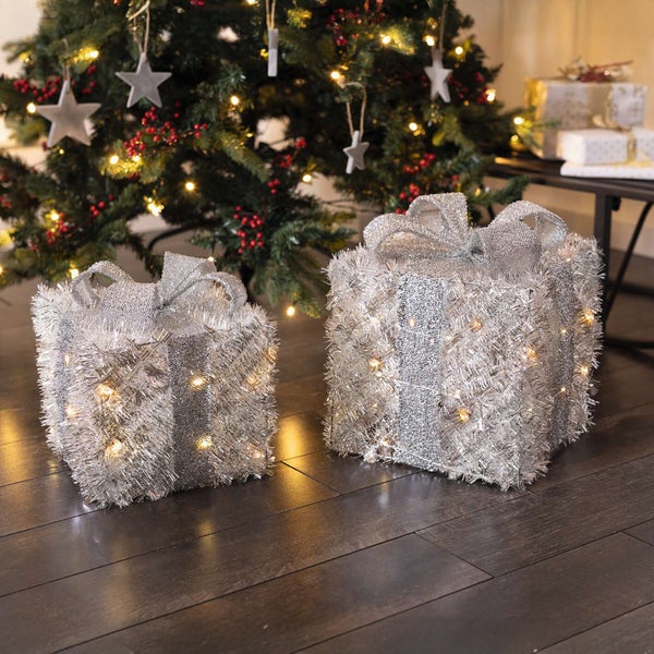 Zwei silberfarbene Geschenkverpackungen mit Schleife und Beleuchtung unter einem Weihnachtsbaum
